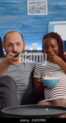 POV von interracial paar mit Schwangerschaft Fernsehen und Popcorn essen. Multiethnische Menschen, die ein Kind erwarten, sitzen auf dem Sofa. Partner, die Spaß haben, während sie die Kamera betrachten Stockfoto