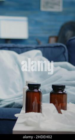 Kranker Mann schläft auf der Couch mit Decke, während er Erkältung und Grippe hat. Person, die wegen einer Viruserkrankung ruht und Flaschen mit Pillen, Medikamenten, einer Tasse Tee und Taschentüchern auf dem Tisch hat. Stockfoto