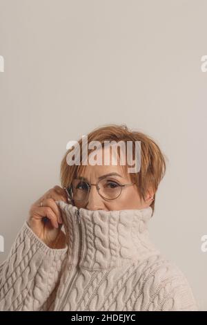 Schöne junge Boomerin mittleren Alters in einer Brille, die in beigen Pullover auf neutralem Hintergrund gewickelt ist. Stockfoto