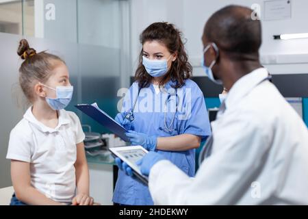 afroamerikanischer Kinderarzt, der die Erfahrung mit Krankheitssymptomen mithilfe einer Tablette analysiert und die Behandlung im Gesundheitswesen mit einem Astant im Krankenhausbüro diskutiert. Ärzteteam mit Gesichtsmaske gegen covid19 Stockfoto