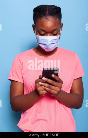 afroamerikanische Studentin mit schützender medizinischer Gesichtsmaske gegen Coronavirus, die im Studio mit blauem Hintergrund ein Smartphone mit SMS-Nachrichten mit Freunden hält. Frau gibt Text ein Stockfoto