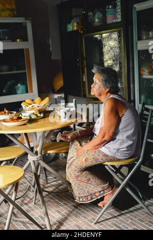 Asiatische alte Großmutter, die frisch gekochte traditionelle thailändische Gerichte serviert und einen Tisch in der rustikalen Küche zu Hause aufgestellt hat. Familienkonzept für die Zubereitung von Abendessen. Stockfoto