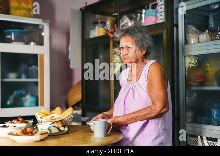 Asiatische alte Großmutter, die frisch gekochte traditionelle thailändische Gerichte serviert und einen Tisch in der rustikalen Küche zu Hause aufgestellt hat. Familienkonzept für die Zubereitung von Abendessen. Stockfoto