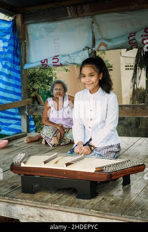 Ein wunderschönes thailändisches Mädchen, das Khim, das traditionelle thailändische Musikinstrument in der Nähe der Großmutter, im Freien auf einer Holzplatte spielt. Freizeit- und Hobbykonzept. Stockfoto