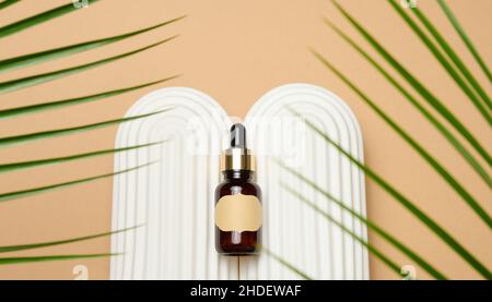 Braune Glasflasche mit Pipette und leerem Papieretikett auf hellbraunem Hintergrund, in der Nähe des Palmblatts. Mockup Hautpflege kosmetisches Produkt Stockfoto