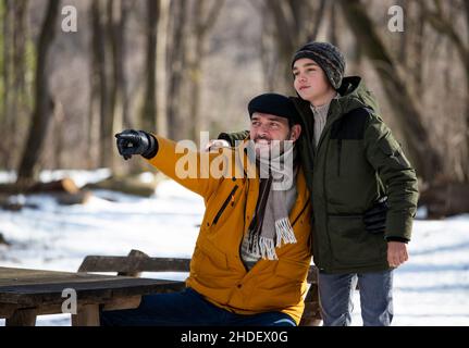 Vater und Sohn sitzen auf der Bank im Wald auf Schnee, während der Winterferien und blicken nach vorne Stockfoto