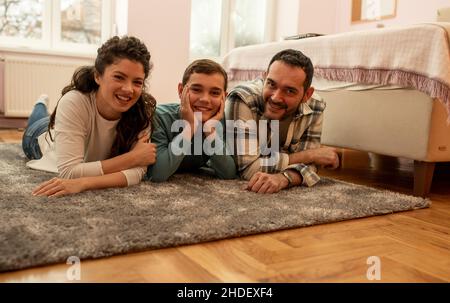 Porträt einer glücklichen dreiköpfigen Familie, Vater, Mutter und Sohn, die auf dem Boden neben dem Bett im Schlafzimmer liegen Stockfoto