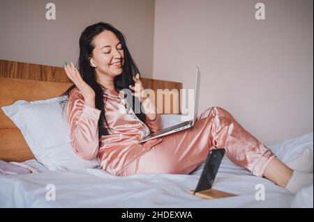 Junge glückliche Brünette Frau mit kabellosen Ohrhörern im Bett in rosa Seide Pyjamas mit Smartphone und Laptop-Computer liegen, haben Online-Video-Chat. Blogger sendet Live-Morgenstream. Stockfoto