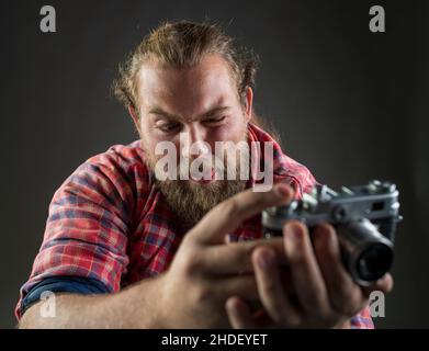 Junger bärtiger Mann, der witzige Gesichtsausdrücke macht, während er ein Foto mit einer altmodischen Kamera macht. Stockfoto