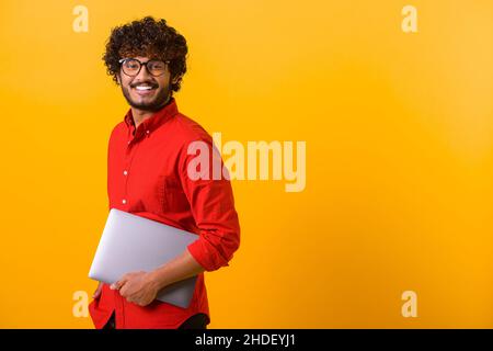 Die Taille nach oben Porträtansicht des lächelnden positiven indischen Mannes, der Laptop in den Händen hält und irgendwo hingeht. Innenaufnahme des Studios isoliert auf orangefarbenem Hintergrund Stockfoto