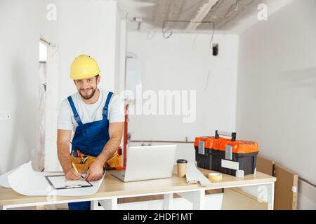 Professioneller Reparaturmann in Uniform und Harthut lächelt der Kamera zu, während er Details eines Auftrags aufschreibt und sich auf Renovierungsarbeiten vorbereitet Stockfoto