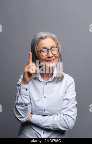 Die vertikale Aufnahme der lächelnden reifen grauhaarigen Geschäftsfrau, isoliert auf grauem Hintergrund, zeigt den Finger nach oben. Dame, die auf etwas achtet Stockfoto