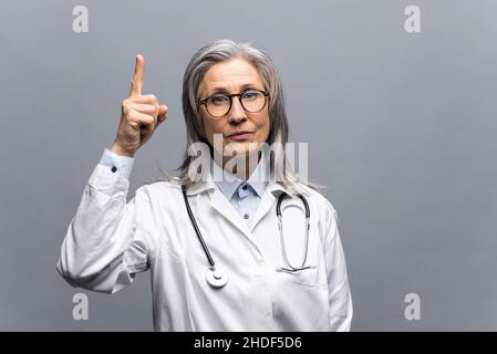 Lächelnde reife grauhaarige Ärztin in weißem medizinischen Gewand, mit Stethoskop isoliert auf grauem Hintergrund zeigt den Finger nach oben. MD achtet auf Krankenversicherung und Krankenversicherung Stockfoto