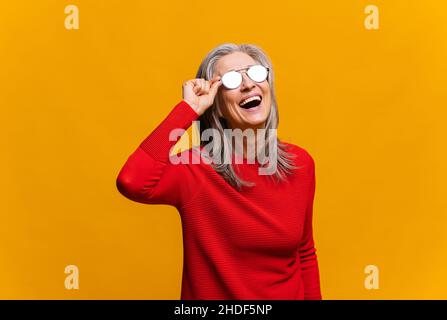Charmante ältere Frau in Sonnenbrille lachend toothy. Elegante, sorglose, reife, moderne, grauhaarige Dame im roten Pullover, die auf gelbem Hintergrund laut aufsteht und lacht. Happy Lifestyle-Konzept Stockfoto