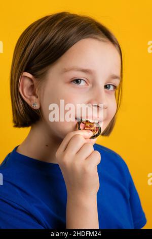 Kleines Mädchen 8-10 Jahre alt in Basic Blue T-Shirt beißen Golden Physical Bitcoin auf gelbem Hintergrund. Kinder Finanzielle Kompetenz. Kryptowährung und Blockchain-Konzept. Stockfoto