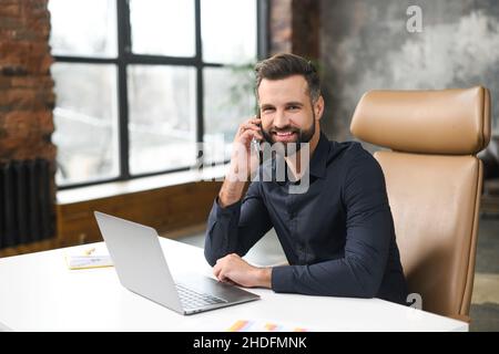 Lächelnder kaukasischer Kerl, der am Schreibtisch sitzt und einen trendigen Laptop für seine Arbeit benutzt. Der junge Mann spricht auf dem Smartphone und lächelt zur Kamera, während er mit Kunden oder Kollegen telefoniert Stockfoto