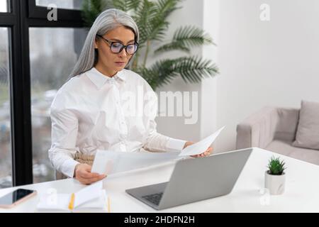 Selbstbewusste ältere Geschäftsfrau macht Schreibarbeiten, während sie mit einem Laptop am Tisch im Büro sitzt. Fokussierter leitender Mitarbeiter, der Dokumente vorbereitet und Rechnungen während der Arbeit überprüft Stockfoto