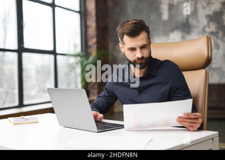 Besorgter kaukasischer Mann, der die Korrespondenz durchschaut. Mann, der Dokumente in den Händen hielt, wurde per Post in Ordnung. Konzentrierter Mann, der mit einem Laptop am Schreibtisch im modernen Büro sitzt Stockfoto