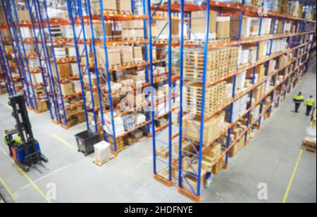 Verwischen Sie den Lagerhintergrund. Ansicht von oben, wie Lagerarbeiter Waren umziehen und Lagerbestände im Gang zwischen Reihen hoher Regale voller verpackter Boxen zählen Stockfoto