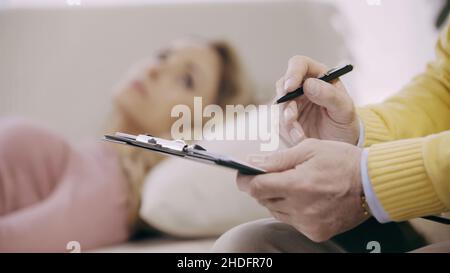 Nahaufnahme eines reifen Psychotherapeuten mit Klemmbrett und Stift in der Nähe eines verschwommenen Patienten Stockfoto