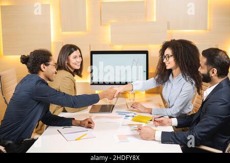 Multirassisches Team, das am Tisch sitzt, lächelt und ihre Hände an einer Stelle zusammenhält, während es Einheit und Teamgeist spürt. Konzept des Arbeitsprozesses Stockfoto