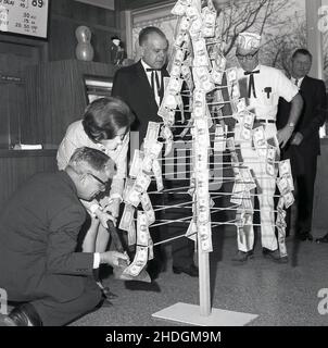 1960s, Historcai, in einem Fast-Food-Outlet von Kentury Fried Chicken (KFC), eine Dame mit einer Axt, die vorgibt, einen Geldbaum abzuhauen, ein Metallständer mit Dollarscheinen, die an den Masten befestigt sind, USA. Stockfoto