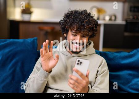 Aufgeregter junger Inder mit Hoodie auf der Couch sitzend und mit Smartphone für Videoverbindung, winkende Hand, Gruß an Freund oder Familie auf der Ferne, ethnischer lockiger Kerl, der Videoanruf abgibt Stockfoto