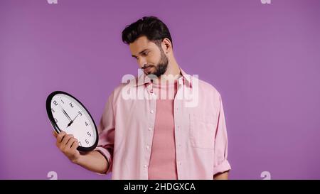 Unzufriedener junger Mann, der die Uhr isoliert auf Purpur ansah Stockfoto