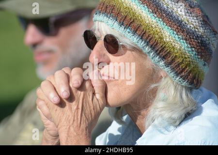 Die ältere, grauende Frau aus den 60ern mit Sonnenbrille und handgestrickter, warmer Strickmütze, die sich Sorgen macht, sich aber darüber freut, wie es ihrem Enkelkind im Fußballspiel geht Stockfoto