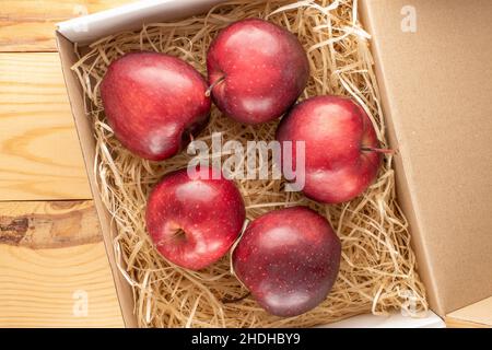 Mehrere reife rote Äpfel in einer Schachtel mit Holzschnitzeln auf einem Holztisch, Nahaufnahme, Draufsicht. Stockfoto