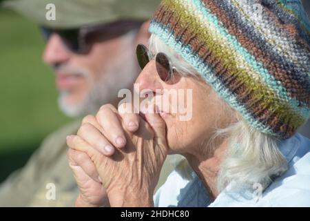 Die ältere, grauende Frau aus den 60ern mit Sonnenbrille und handgestrickter, warmer Strickmütze, die sich Sorgen macht, sich aber darüber freut, wie es ihrem Enkelkind im Fußballspiel geht Stockfoto