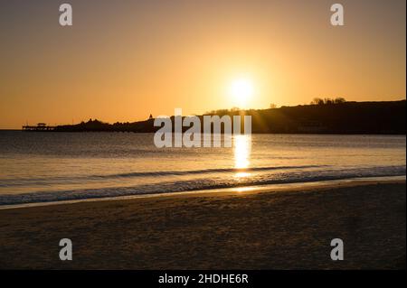 Sonnenaufgang in Swanage mit Blick über die Bucht mit dem Swanage Pier, dem Gipfel der Hügel und dem Wellington Clock Tower in Silhouette mit klarem Himmel und Sandstrand. Stockfoto