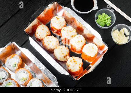 Sushi-Brötchen zum Mitnehmen in Behältern, philadelphia-Brötchen und gebackene Garnelenrollen auf schwarzem Holztisch im Hintergrund Stockfoto