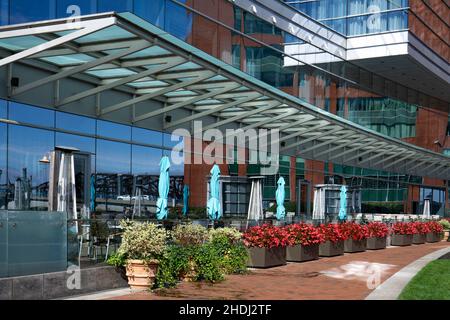 Straßencafé mit Heizlampen und Sonnenschirmen neben dem Glaswandgebäude. Eingetopfte rote Blumen als Dekoration Stockfoto