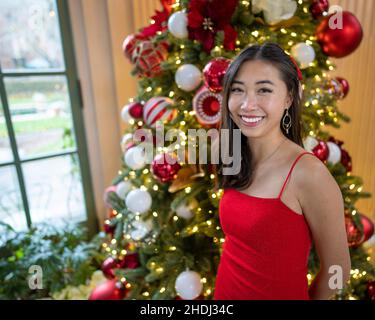 Junge ostasiatische Frau in einem kurzen roten Kleid, die vor einem exquisit dekorierten Weihnachtsbaum steht Stockfoto