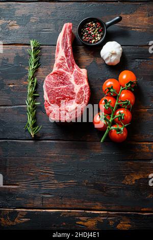Rohes, ungekocktes, schwarzes angus-Rind Ribeye Steak, Bone-Incloseup mit Tomaten, Knoblauchkräutern und Pfefferkörnern, Rosmarin auf hölzernen, dunklen, rustikalen Tischplanken Stockfoto