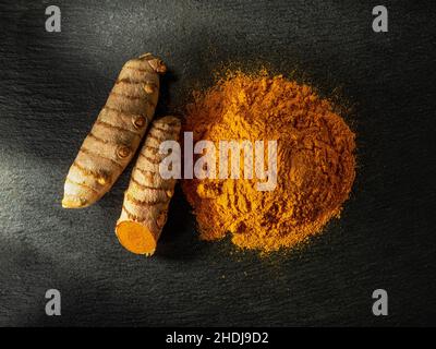 Organisches Kurkuma-Pulver und frische Curcuma-Wurzeln auf einer schwarzen Schieferoberfläche. Rohe und gemahlene Rhizome von Curcuma longa. Indische Gewürze, Zutat für das Indische. Stockfoto