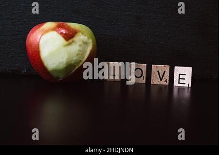 Das Wort Liebe lag mit Holzbuchstaben auf schwarzem Hintergrund mit einem Apfel, in den ein Herz geschnitten wurde Stockfoto