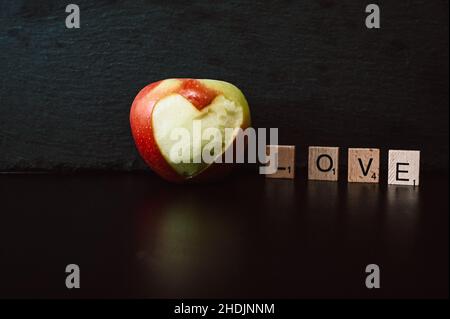 Das Wort Liebe aus Holzbuchstaben auf schwarzem Hintergrund, neben einem Apfel mit ausgeschnittenem Herzen Stockfoto