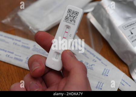 In der Hand wurde ein positiver Covid-19-Lateral-Durchflusstest durchgeführt, der die beiden roten Markierungslinien zeigt. Teil eines Sets mit unterschiedlichen Hintergründen und Testwerkzeugen. Stockfoto