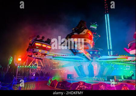 Menschen, die nachts auf einem sich drehenden Messegelände mit Raucheffekt fahren, der leuchtende Farben erzeugt Stockfoto