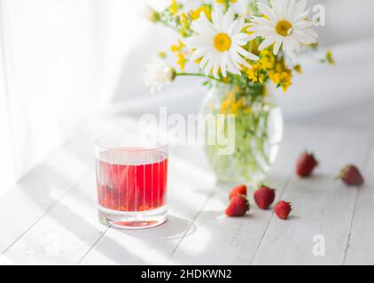 Gänseblümchen und gelbe Blumen in einer klaren Vase auf dem Tisch und ein Glas Beerengetränk Stockfoto