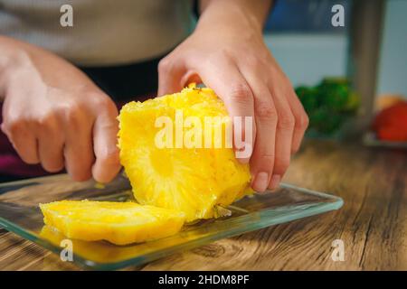 Nahaufnahme der Hände einer jungen Frau, die mit einem Kochmesser auf einem Holztisch in einer Küche eine saftige Ananas schneidet. Stockfoto