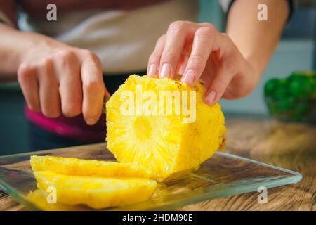 Nahaufnahme der Hände einer jungen Frau, die mit einem Kochmesser auf einem Holztisch in einer Küche eine saftige Ananas schneidet. Stockfoto