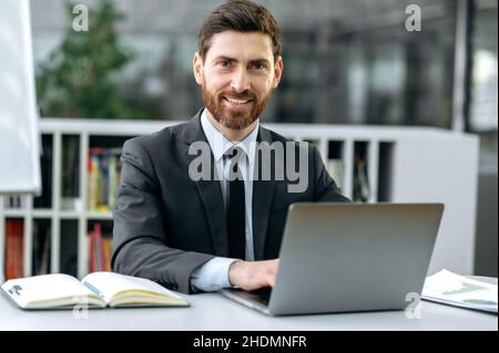 Portrait ein gutaussehender, erfolgreicher kaukasischer bärtiger Geschäftsmann, ceo des Unternehmens, Top-Manager, in formellem Anzug, sitzend am Tisch mit Laptop im modernen Büro, schauend auf die Kamera und lächelnd freundlich Stockfoto