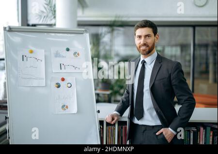 Porträt intelligent attraktiv positiv bärtig kaukasischer Erwachsener Geschäftsmann, Finanzanalyst, Abteilungsleiter, steht in der Nähe des Whiteboards im Büro, Blick auf die Kamera, lächelnd freundlich Stockfoto