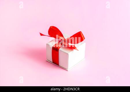 Kleine weiß verpackte Geschenkbox mit leuchtend rotem Schleifband auf hellrosa Hintergrund. Valentinstag, Geburtstag, Muttertag Überraschung pesent. Stockfoto