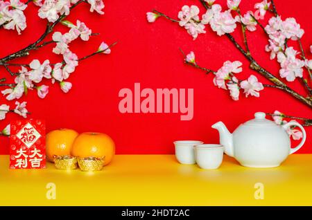 Teeset, Orangen, Ingots und rote Umschlagschachtel oder ang bao (Wort bedeutet großes Glück) mit Pfirsichblüte auf gelbem und rotem Glitterpapier-Hintergrund. Stockfoto