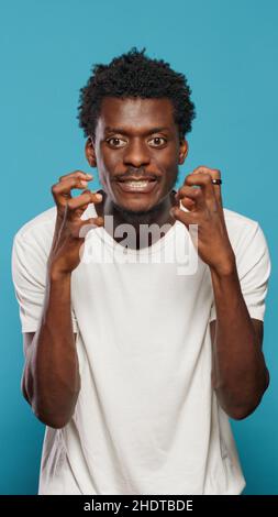 Frustrierter Erwachsener, der sich im Studio wütend fühlt und sich mit den Fäusten festhält. Unzufriedener Mensch, der wegen Wut und Frustration Zähne zeigt und auf blauem Hintergrund steht. Porträt eines aggressiven Mannes Stockfoto