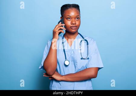 afroamerikanische Krankenschwester, die am Telefon telefoniert und mit dem Therapeuten, der an der Gesundheitsbehandlung arbeitet, über die Erfahrung bei der Behandlung von Krankheiten spricht, im Studio mit blauem Hintergrund. Medizinkonzept Stockfoto